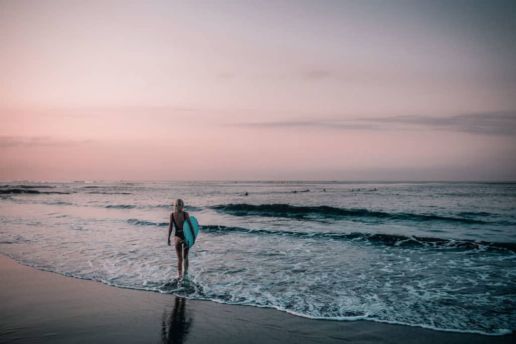 Surfer style girl standing on beach with surfboard at sunset