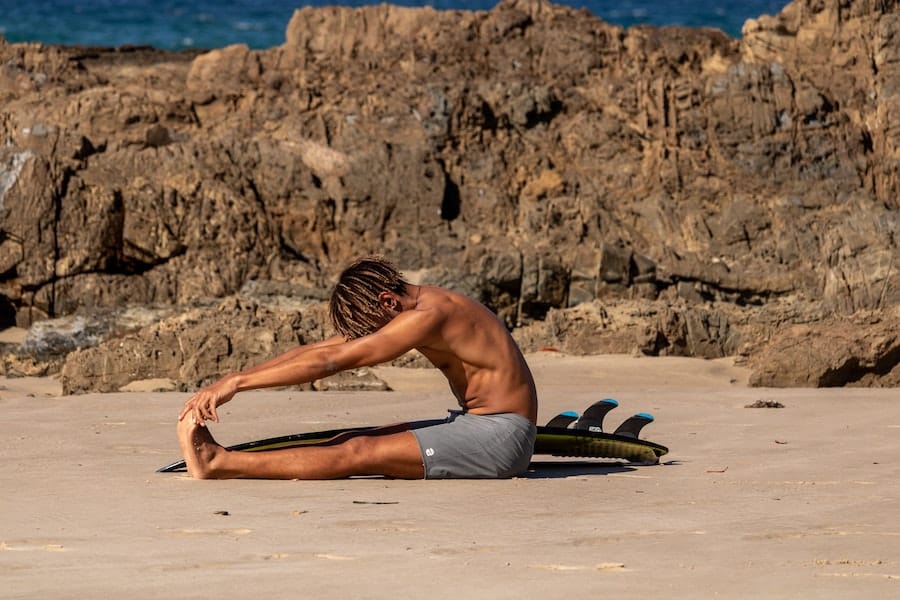Man stretching on the beach with his surfboard beside him