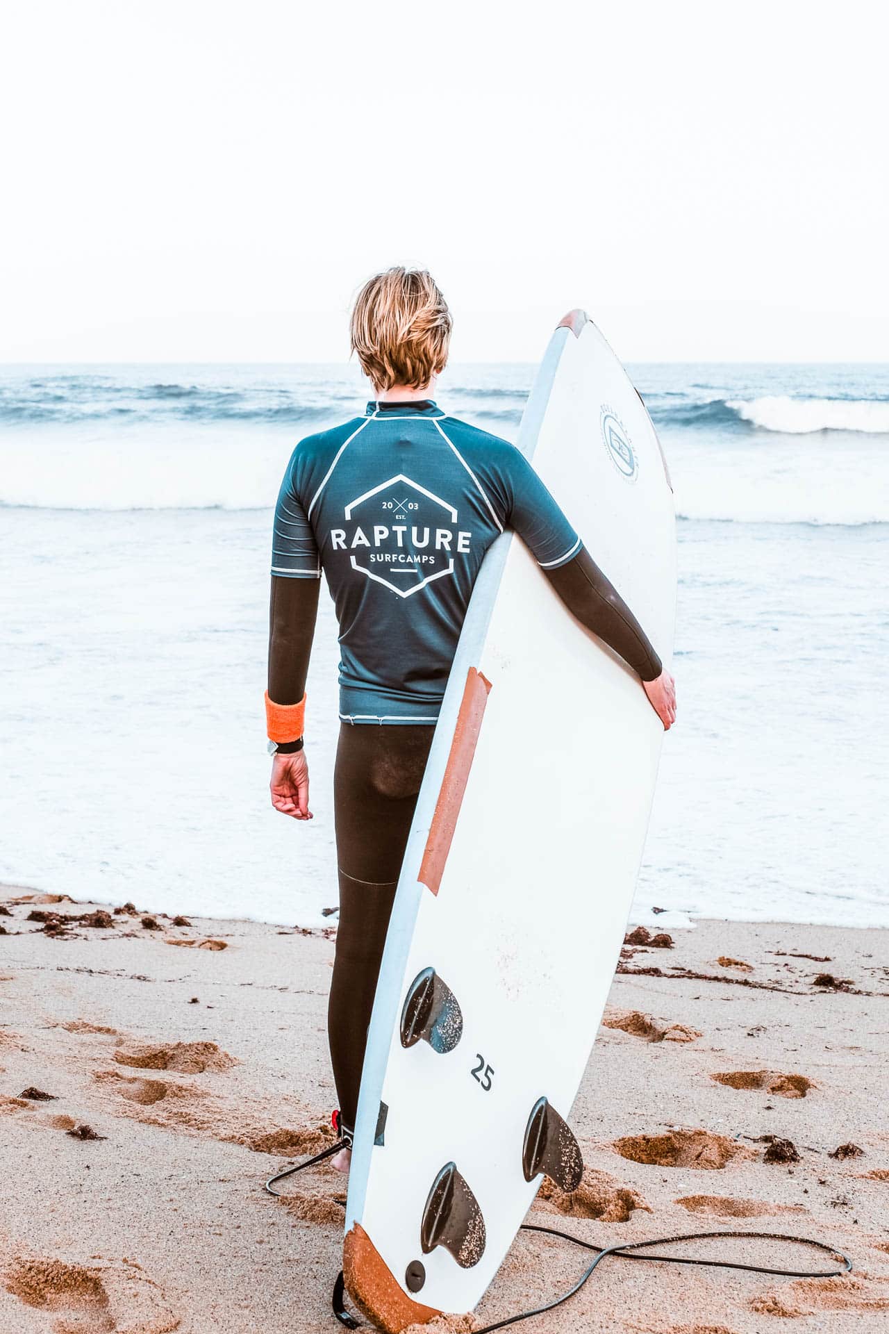 Person wearing Rapture Surfcamps Surf wear holding a surfboard and facing the ocean.