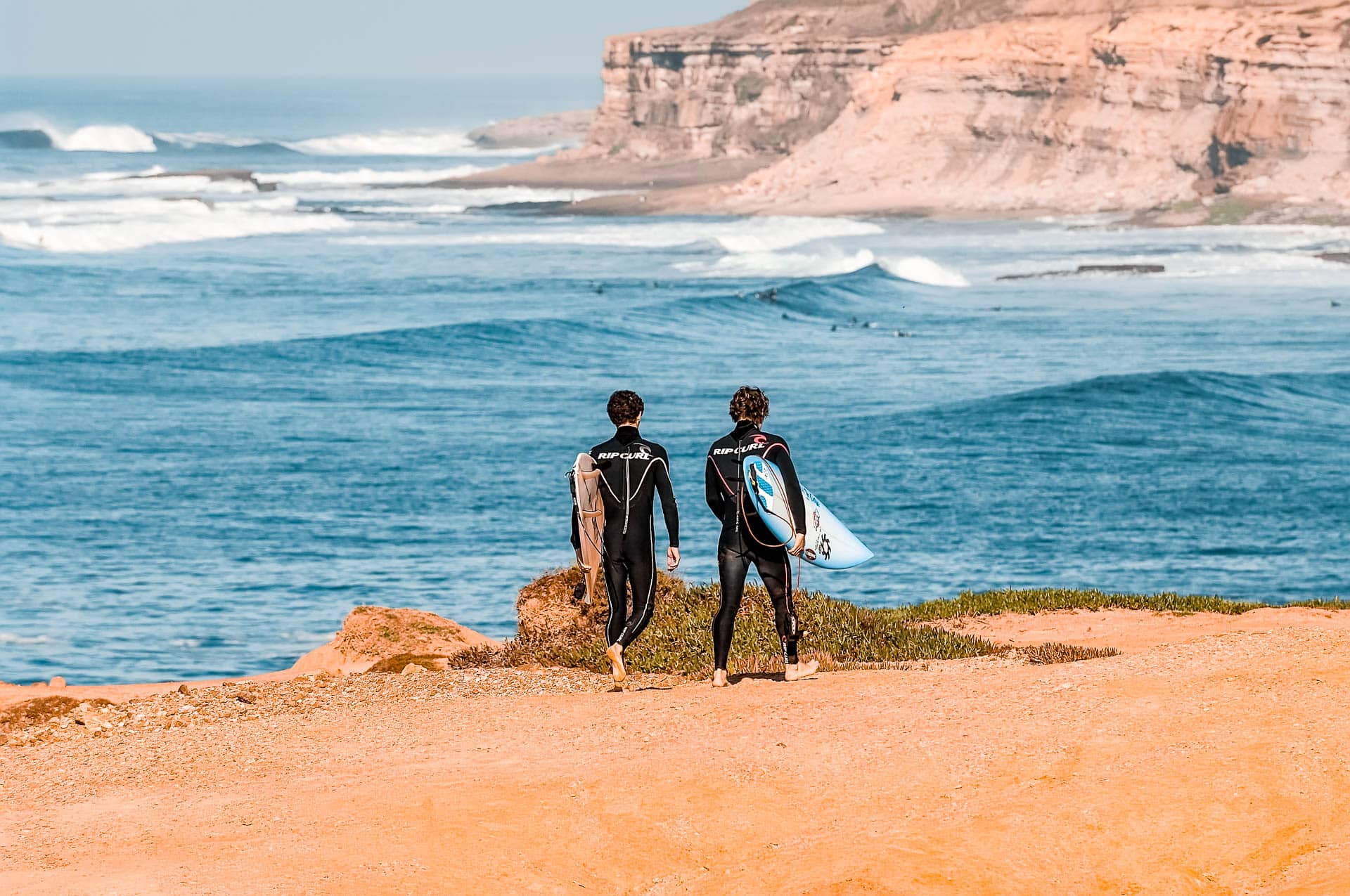 Two surfers in wetsuit walking towards the beach with surfboards in their hand.