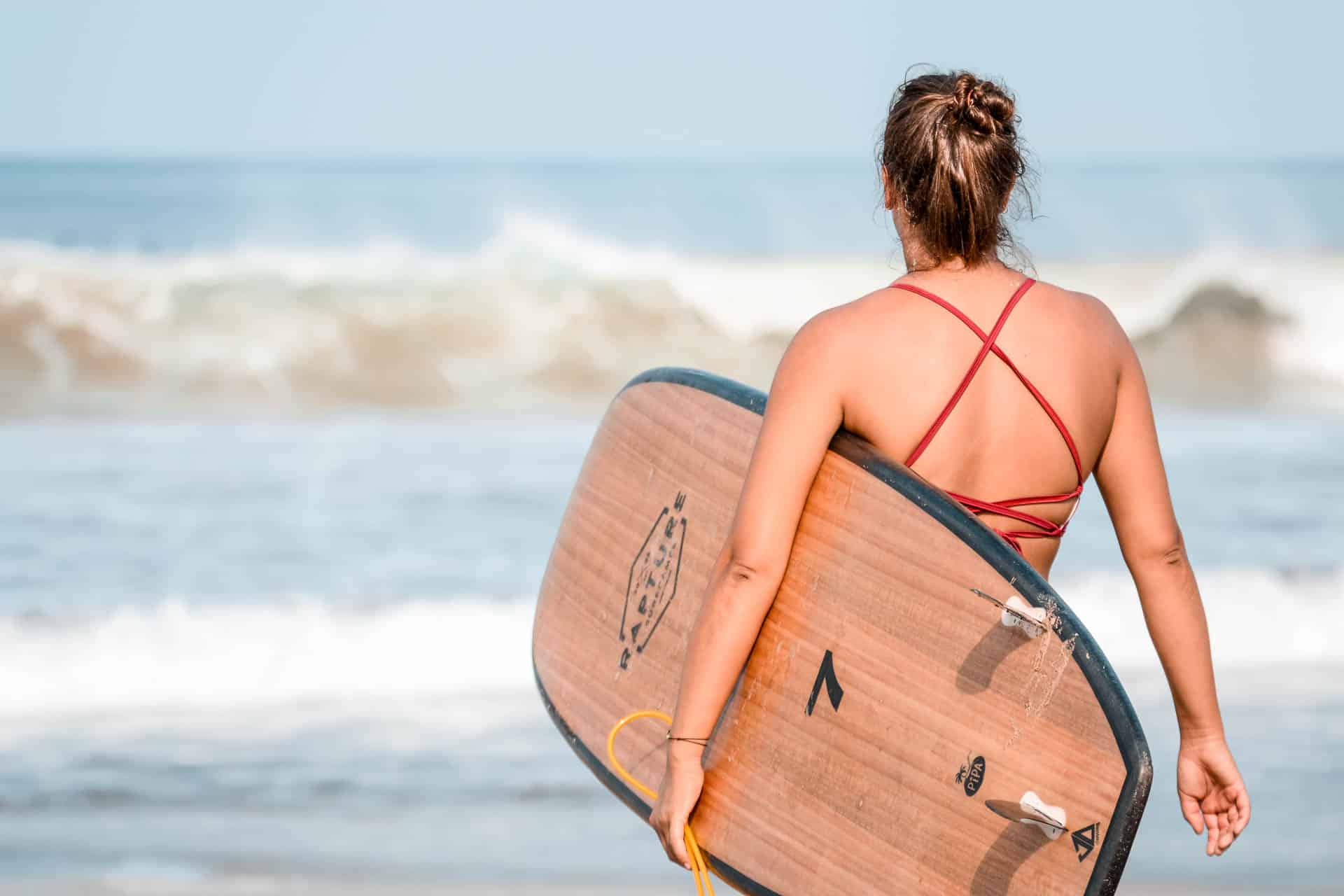 A girl in orange bikini looking at the ocean with a brown surfboard in arm.