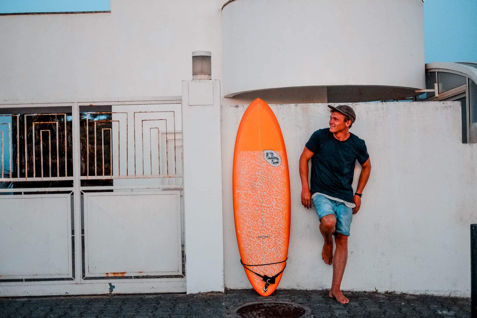 A person leaning against the wall posing with an orange surfboard.