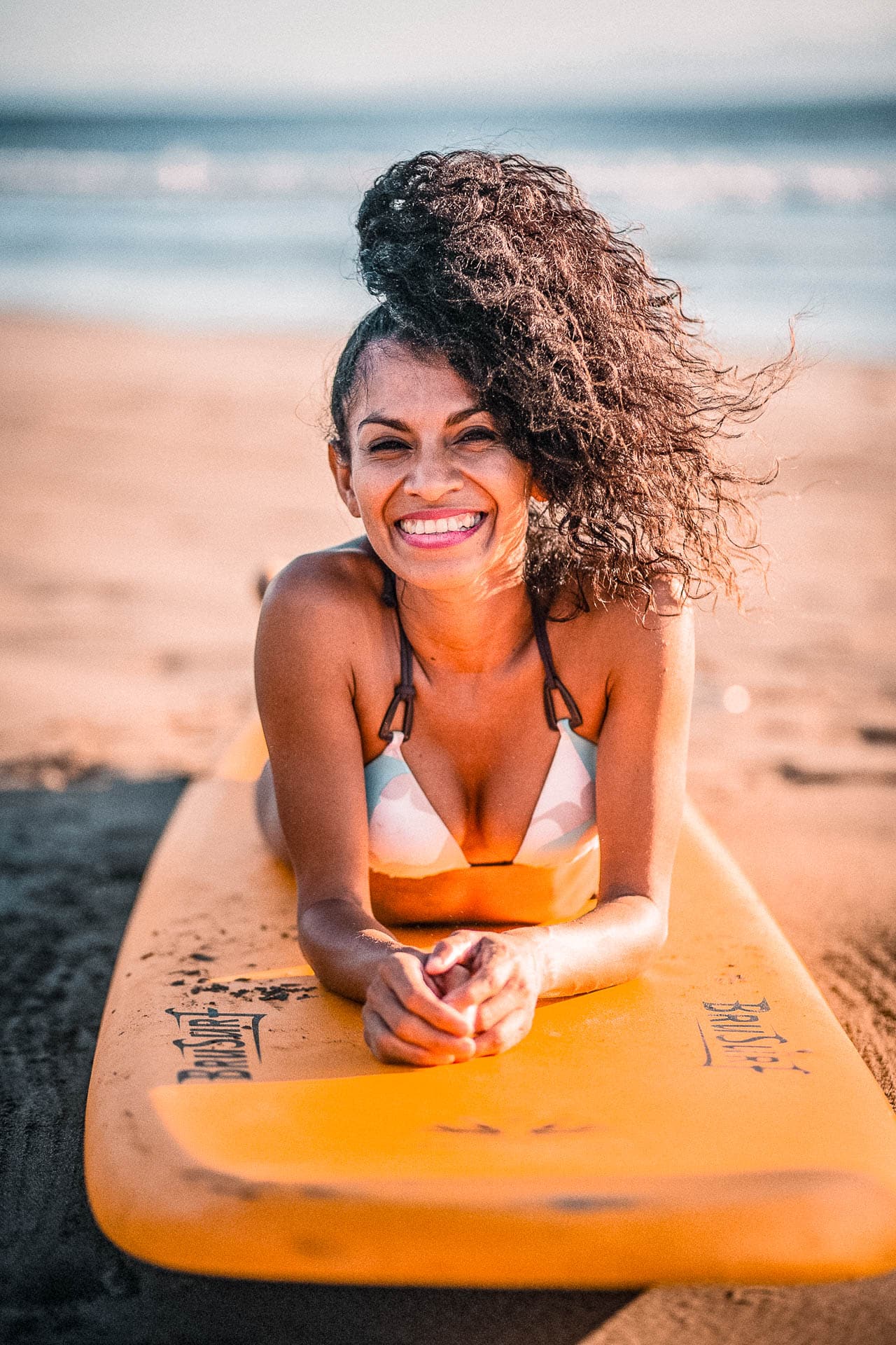 A person with curly long hair tied in a pony posing on a yellow surfboard