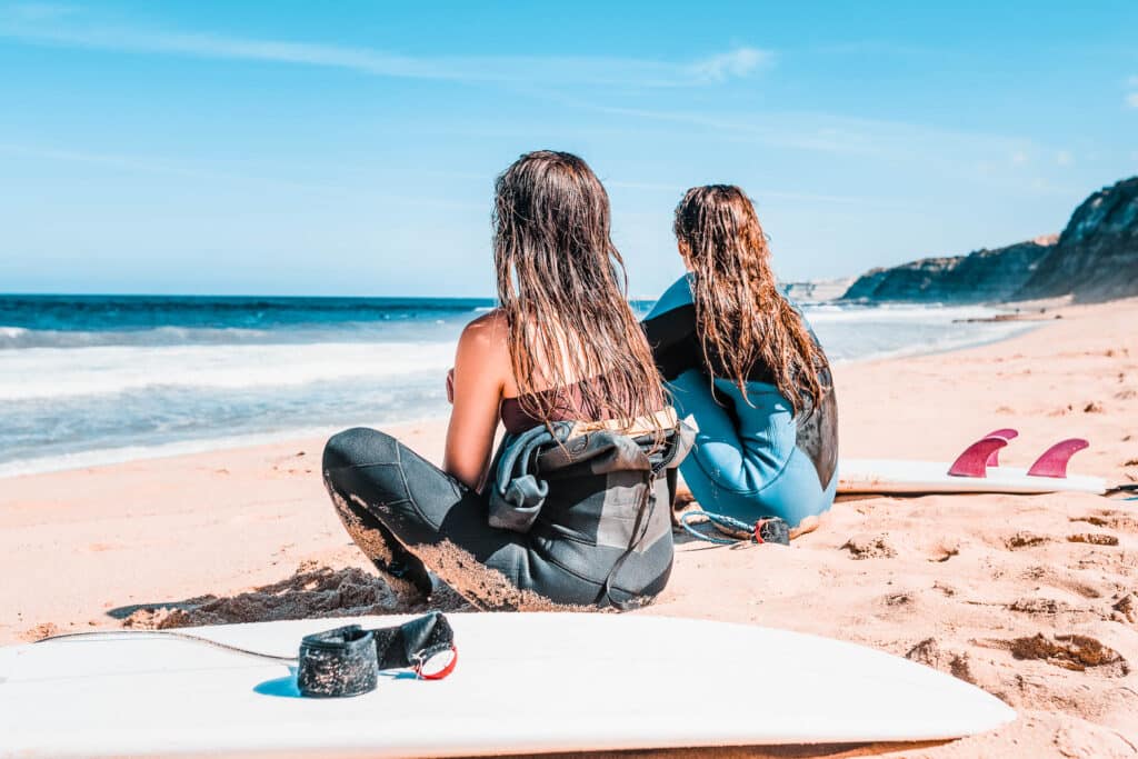 Two people in wetsuit sitting on the beach with their surfboards looking at the ocean.