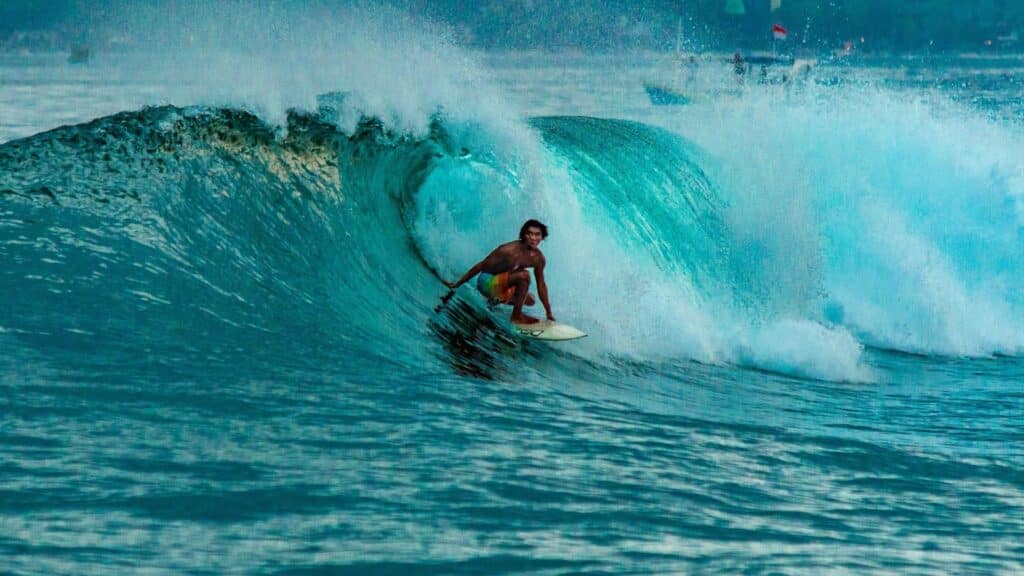 A surfer riding a vibrant turquoise wave, leaning into the curve of the water with skill and focus. The ocean spray creates a dynamic and energetic atmosphere, with the background showing a faint outline of distant figures and flags